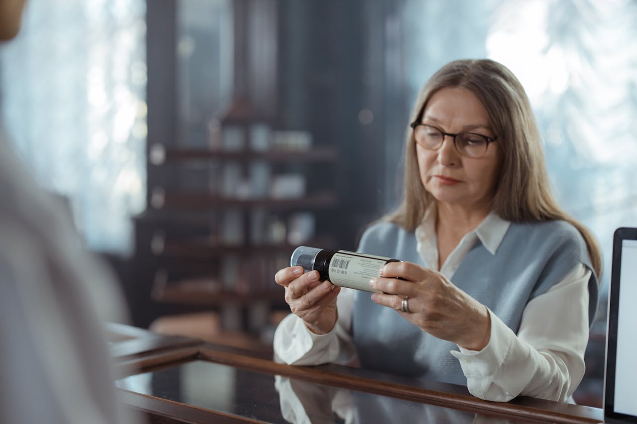 Mastering the First Impression: Your intriguing post title goes here A senior woman with eyeglasses reviews a medicine bottle at a pharmacy counter indoors.